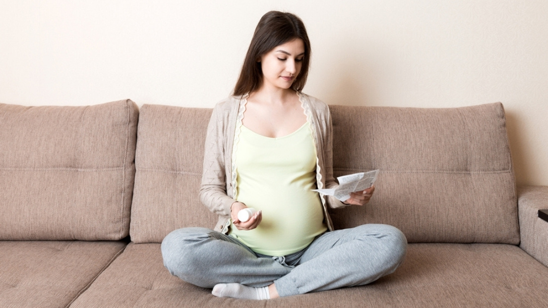 Trudnica sedi na kauču i čita uputstvo uz preparat u ruci, proveravajući koje aktivne supstance u gelu za hemoroide su bezbedne za primenu tokom trudnoće
