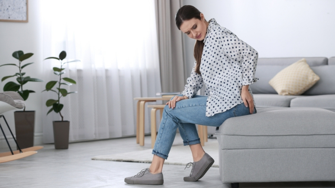 Trudnica sedi na ivici kreveta i drži se za donji deo leđa i karlicu, što ukazuje na nelagodnost i bol povezane sa hemoroidima u trudnoći