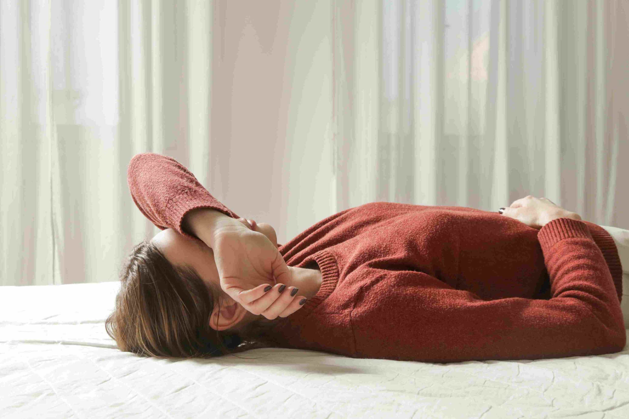 Osoba leži na krevetu i prekriva lice rukom, deluje iscrpljeno i zabrinuto